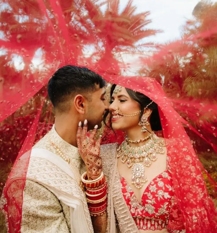 Bride and groom posing under a dupatta in a romantic Indian wedding photoshoot